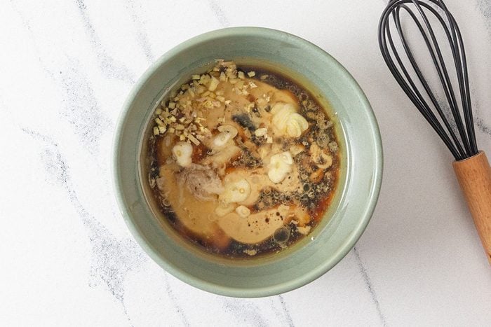 Overhead shot of a green bowl filled with sauce ingredients, including soy sauce, minced garlic, and ginger, placed on a white surface beside a whisk with a wooden handle;