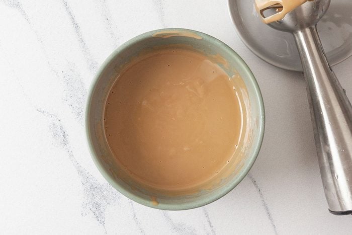 Overhead shot of a green bowl filled with smooth, light brown sauce, set on a white marble surface beside an immersion blender with sauce residue on its blade;