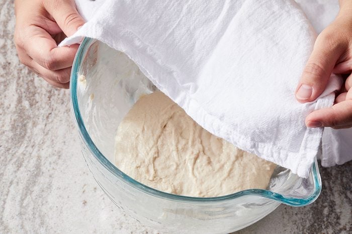 Overhead shot of a person lifting a white cloth to reveal rising dough in a glass mixing bowl on a light-colored surface.