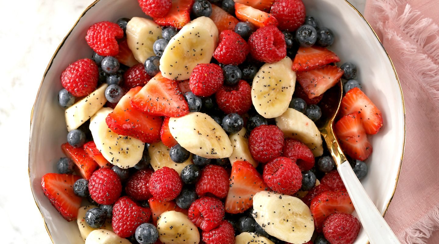 A bowl filled with sliced bananas, strawberries, raspberries, and blueberries, lightly sprinkled with poppy seeds, with a spoon on the side.