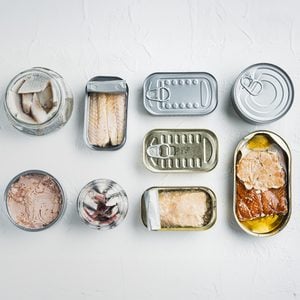 An assortment of opened canned seafood, including fish fillets and chunks, arranged neatly in rows on a white surface. Various can sizes and shapes are visible, showing the variety of preserved seafood inside.